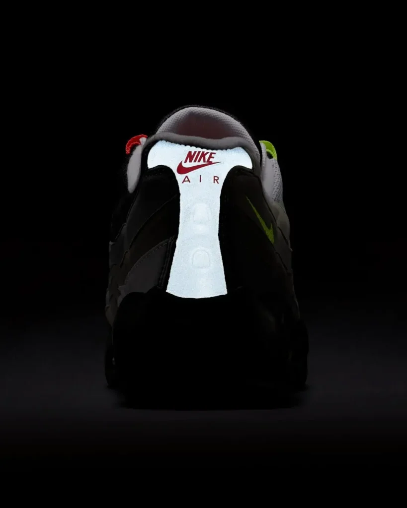 Rear view of a Nike Air Max 95 showing reflective white heel panel with red Nike Air branding, dark upper, and neon green mini Swoosh against a black background