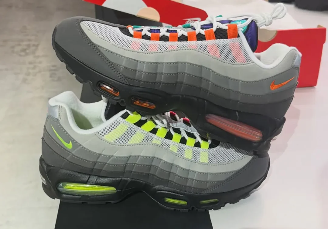 Side view of the Nike Air Max 95 'Greedy' Big Bubble with one shoe stacked atop the other, showcasing mismatched neon lace loops, grey gradient suede panels, white mesh, orange and volt mini Swooshes, and contrasting red and green visible Air units.