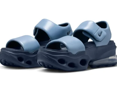 Pair of Nike Air Max platform sandals shown in a three-quarter front view against a white background. The design combines light blue padded forefoot and adjustable ankle straps with a dark navy sculpted foam midsole featuring oval cutout details and a visible full-length Air unit in the heel. Small black Swoosh logos appear on the ankle straps, highlighting the sandal's futuristic blend of comfort and Air Max cushion