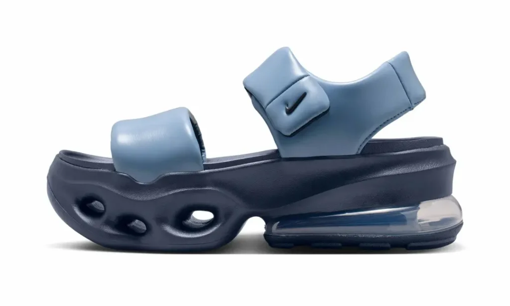 Lateral side profile of the Nike Air Max platform sandal in a blue colorway. The image emphasizes the padded adjustable ankle strap with a black Swoosh logo, wide forefoot strap, sculpted navy foam sole with aerodynamic cutout openings, and the large transparent Air Max heel unit that provides elevated cushion