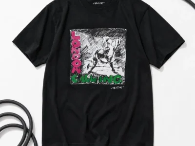Black Tee laid flat on white, with a coiled instrument cable looped around it. Chest graphic is a black-and-white sketch-style illustration of a figure swinging an object, with "LONDON" in pink letters vertically down the left and "CALLING" in green across the bottom.