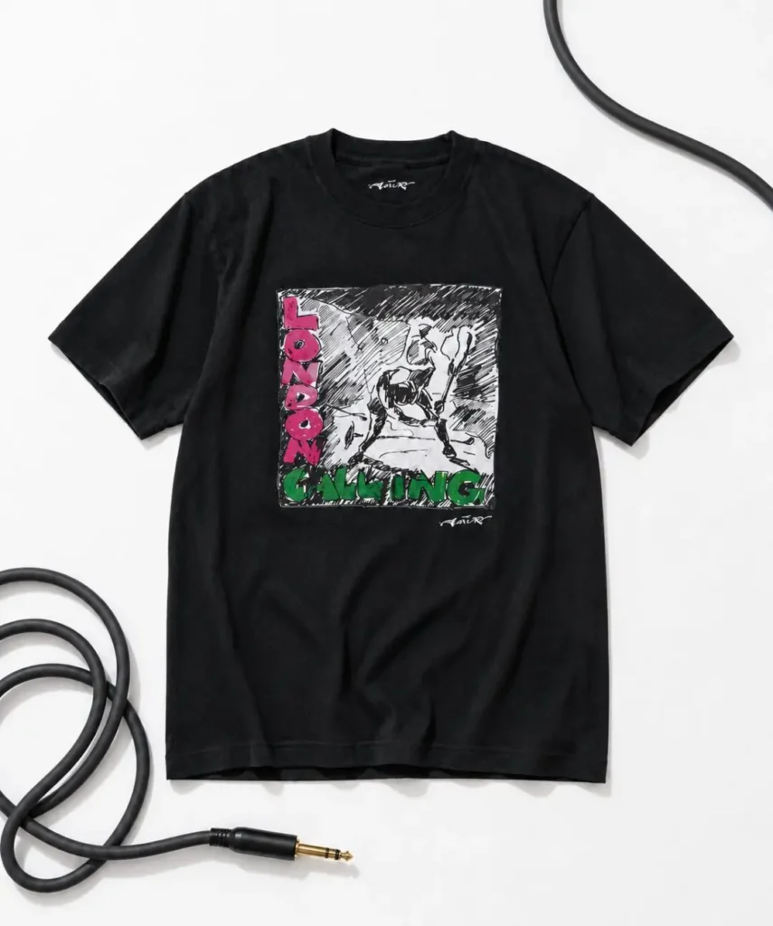Black Tee laid flat on white, with a coiled instrument cable looped around it. Chest graphic is a black-and-white sketch-style illustration of a figure swinging an object, with "LONDON" in pink letters vertically down the left and "CALLING" in green across the bottom.