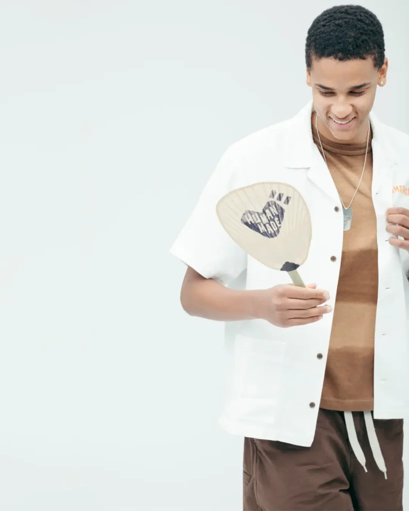 Studio portrait of a smiling model in a white Human Made camp shirt over a brown striped tee while holding a branded Japanese fan