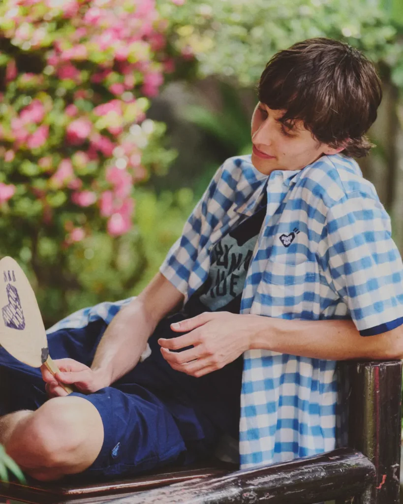 A model lounges on a wooden railing in a blue gingham Human Made overshirt while holding a branded Japanese uchiwa fan beside blooming flower