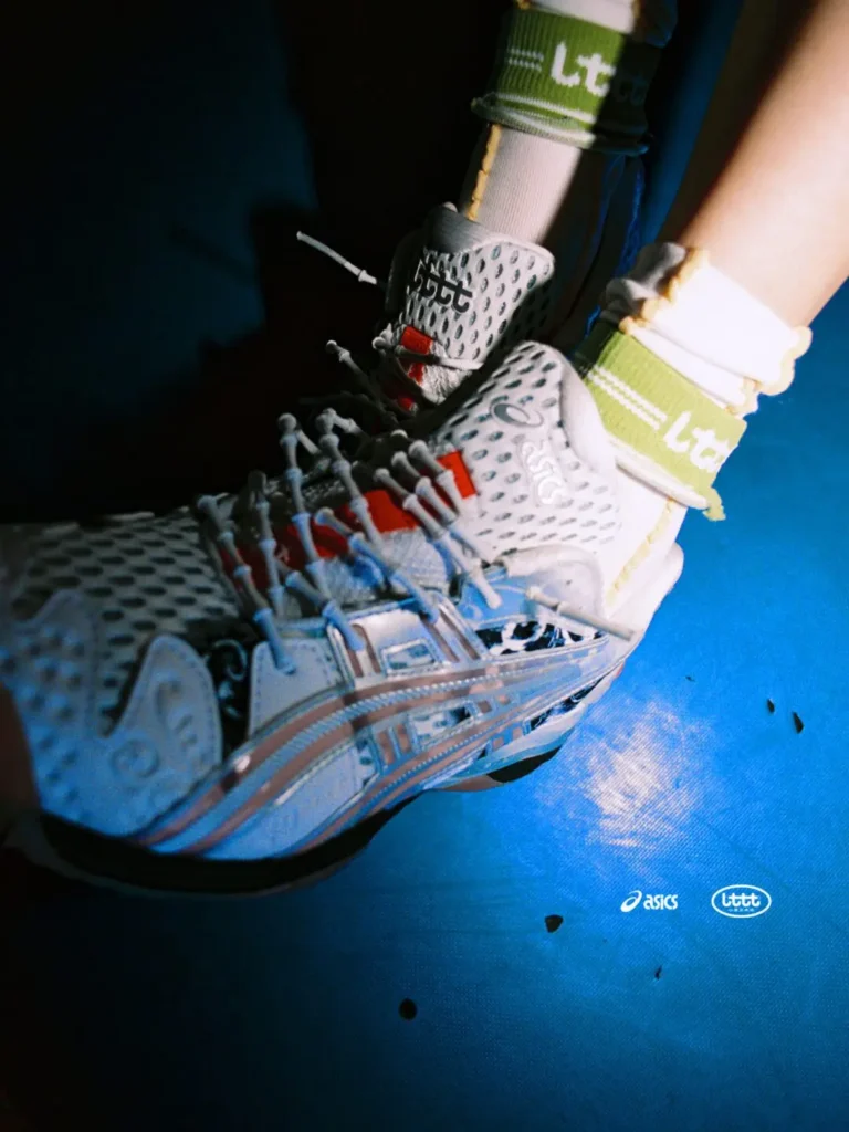 Close-up of the white ASICS x Lttt table tennis shoe worn on court, highlighting the perforated mesh upper, translucent side cage, gum outsole, green secondary laces, and red tongue detail under dramatic light