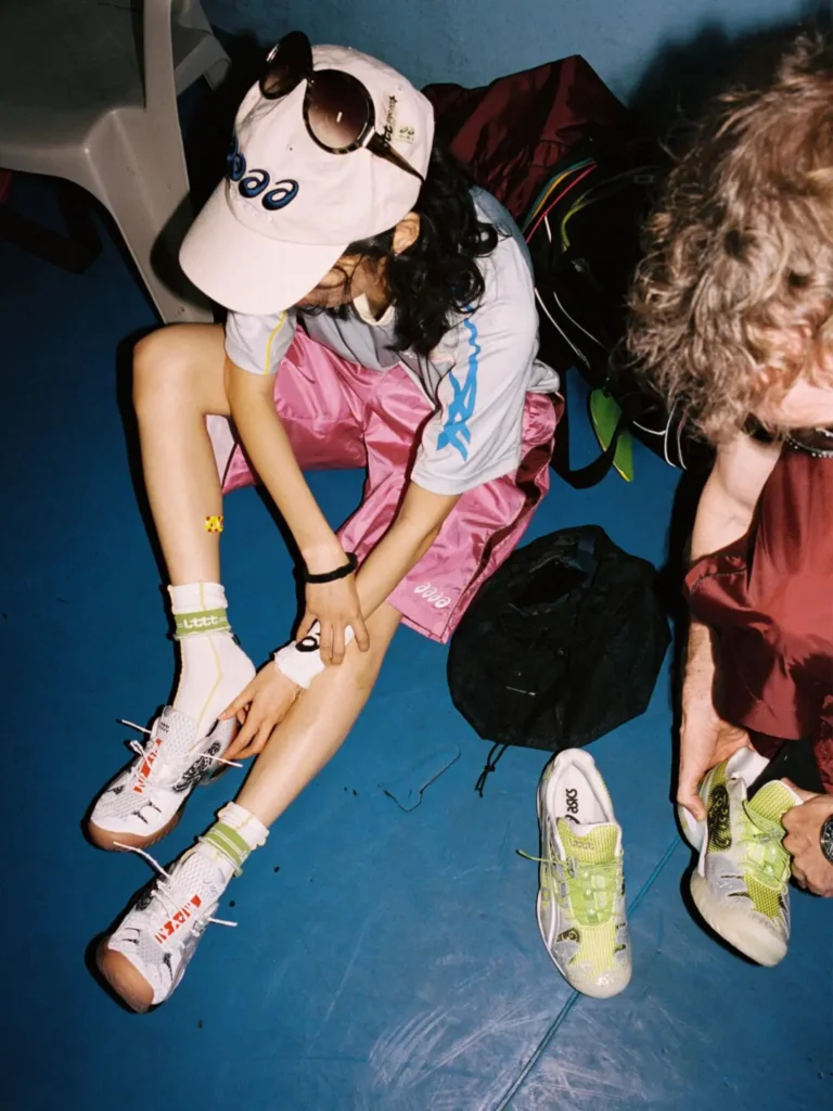 Two table tennis players prepare courtside wearing the ASICS x Lttt collaboration, with one athlete lacing the white pair while another holds the lime green version beside sports bags and training apparel