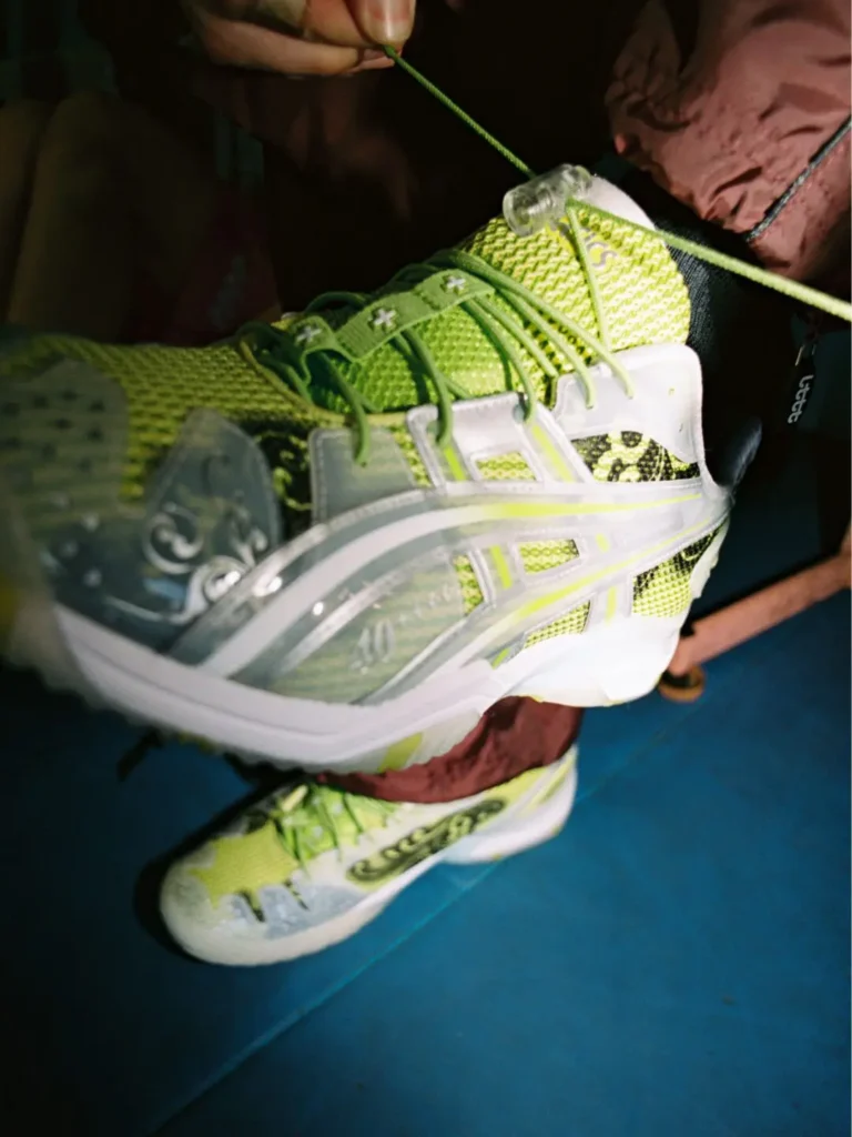 Detailed close-up of the lime green ASICS x Lttt table tennis shoe featuring breathable mesh, translucent ASICS stripes, decorative black side graphics, layered lace system, and semi-translucent toe protection