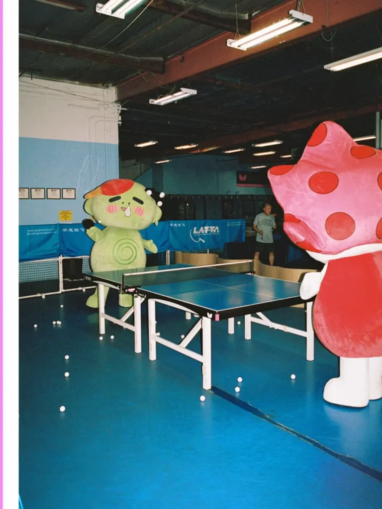 Two oversized mascot characters face each other across a table tennis table inside an indoor sports hall scattered with white ping pong balls, creating a play campaign scene