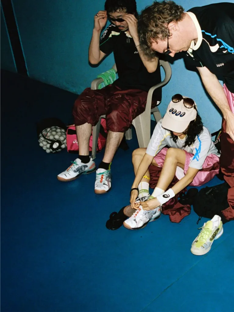 hree players sit and prepare for table tennis action while adjusting footwear and sunglasses, with both ASICS x Lttt collaborative shoe colorways view on the blue indoor court