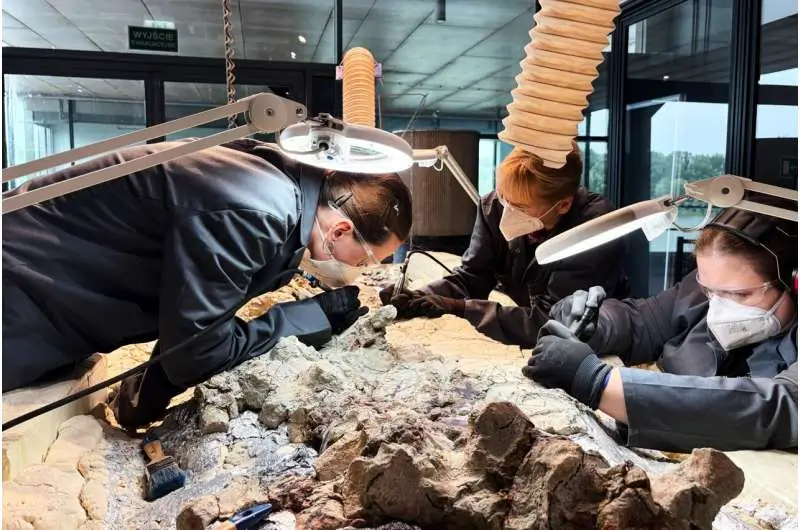 Three paleontology conservators wearing protective masks, gloves, and safety glasses carefully excavate a large dinosaur fossil block under bright magnifying lamps. Precision tools and brushes surround the partially exposed bones inside a modern laboratory with extraction hoses suspended overhead.
