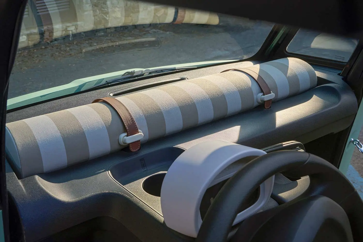 Close-up of the Fiat Topolino Dolcevita dashboard featuring a beige-and-white striped fabric storage roll secured with brown leather straps, minimalist controls, cup holder, and a compact digital instrument cluster.