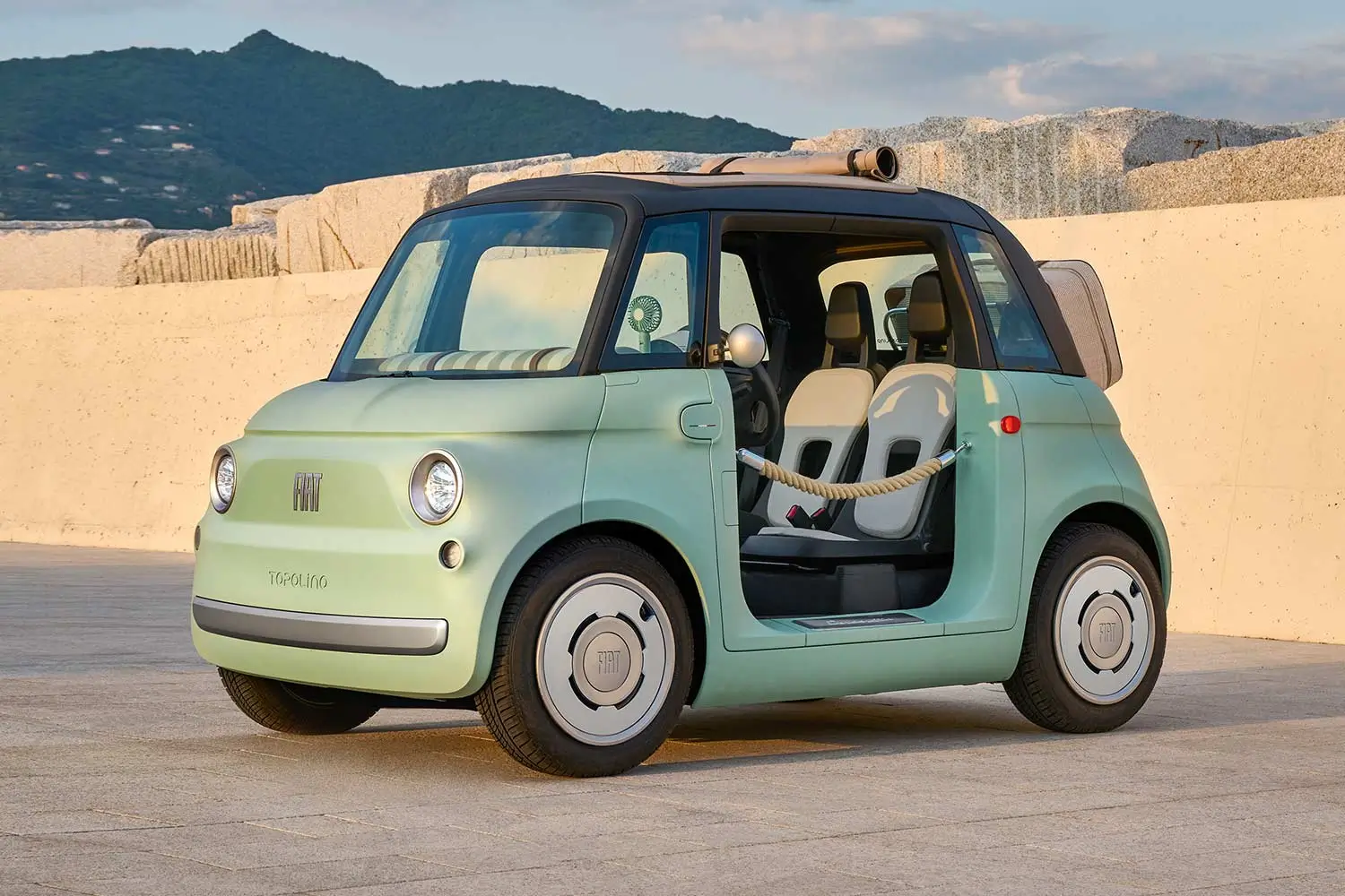 Mint-green Fiat Topolino Dolcevita microcar with an open side entrance, black canvas roof, rope-style door restraint, and minimalist two-seat interior parked in front of a stone wall with mountains in the background.