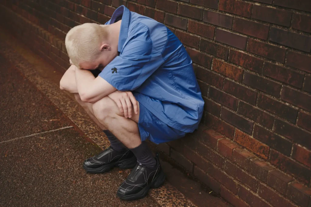A person with short blonde hair wearing a blue shirt and shorts sits on the ground against a brick wall, hunched over with their head resting on their crossed arms, appearing distressed or upset.