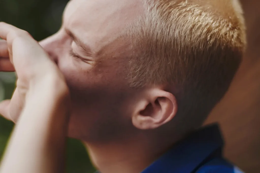 A close-up of a person with short blond hair, eyes closed, and hand covering part of their face, appearing emotional or deep in thought. They are wearing a blue shirt, and the background is blurry.