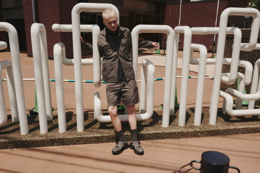 A person with short blond hair wearing a matching brown jacket and shorts stands with arms outstretched, framed by a series of white, curved pipes in an outdoor urban setting.