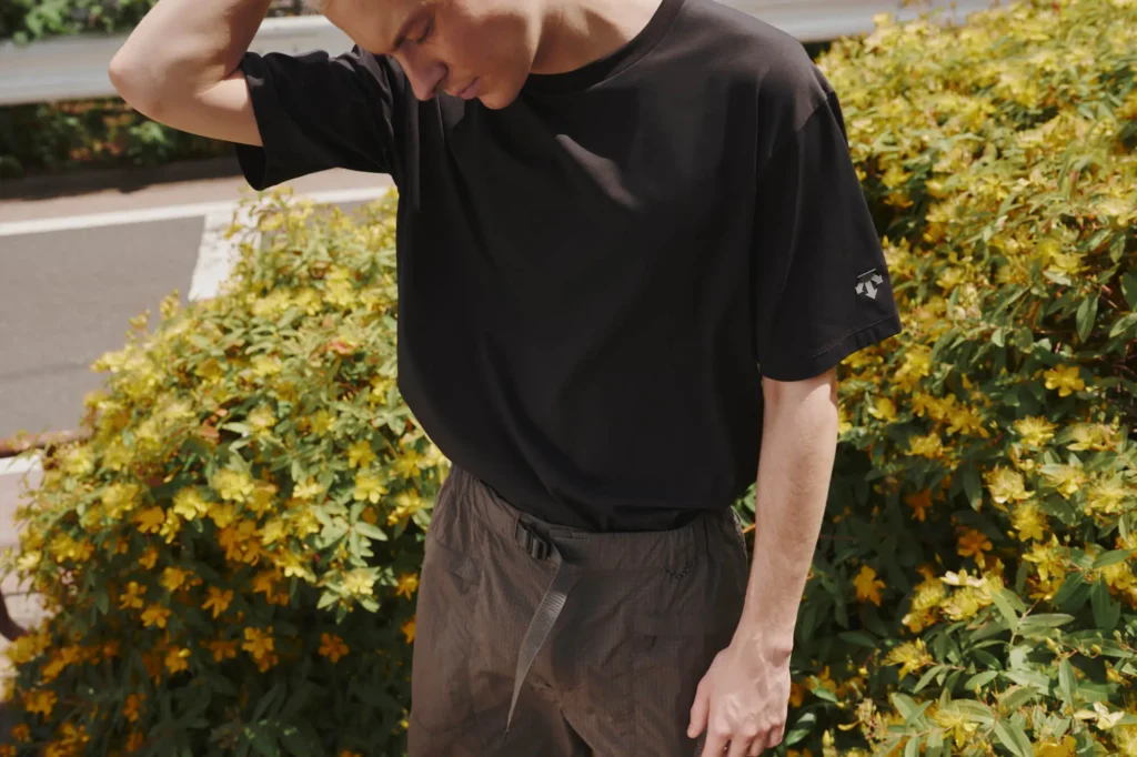 A person stands outdoors in front of green bushes with yellow flowers, wearing a loose black t-shirt and brown shorts, with one hand touching their head and looking down.