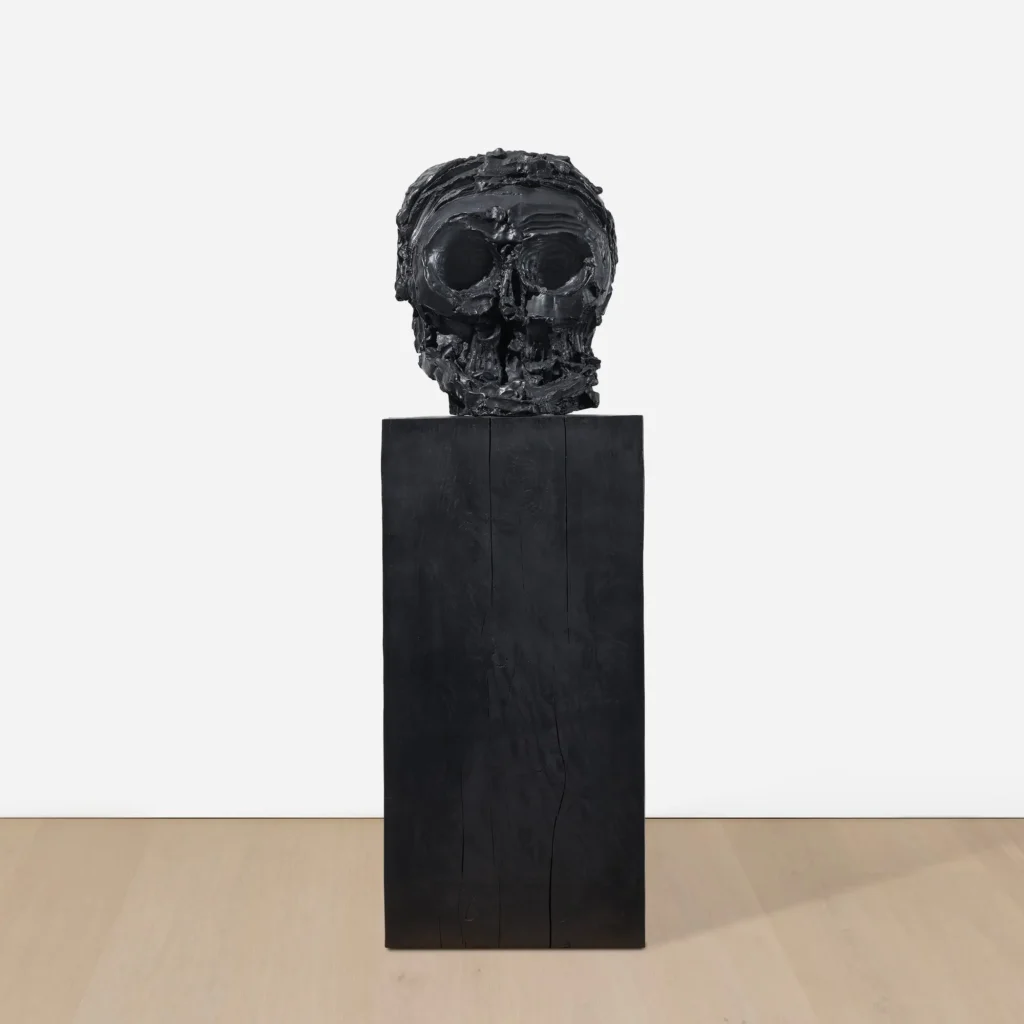 Front view of a heavily textured black skull sculpture with exaggerated eye sockets and fragmented facial features, mounted on a tall matte-black rectangular pedestal in a minimalist gallery