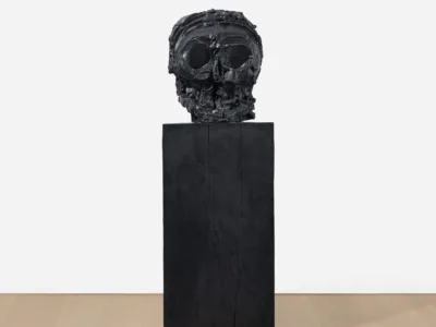 Front view of a heavily textured black skull sculpture with exaggerated eye sockets and fragmented facial features, mounted on a tall matte-black rectangular pedestal in a minimalist gallery