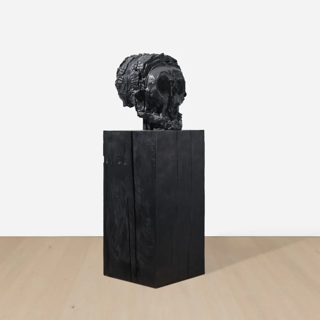 Three-quarter side view of a monochromatic black skull sculpture revealing layered sculptural textures and deep carved surfaces atop a geometric black pedestal