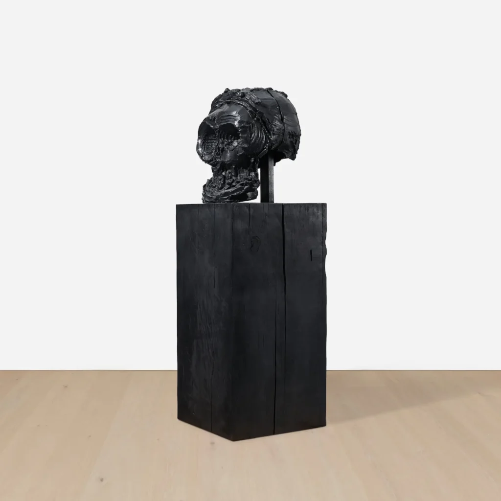 Opposite side profile of a black skull sculpture showing its hollow interior, exposed support column, and rough hand-worked surface on a tall black plinth
