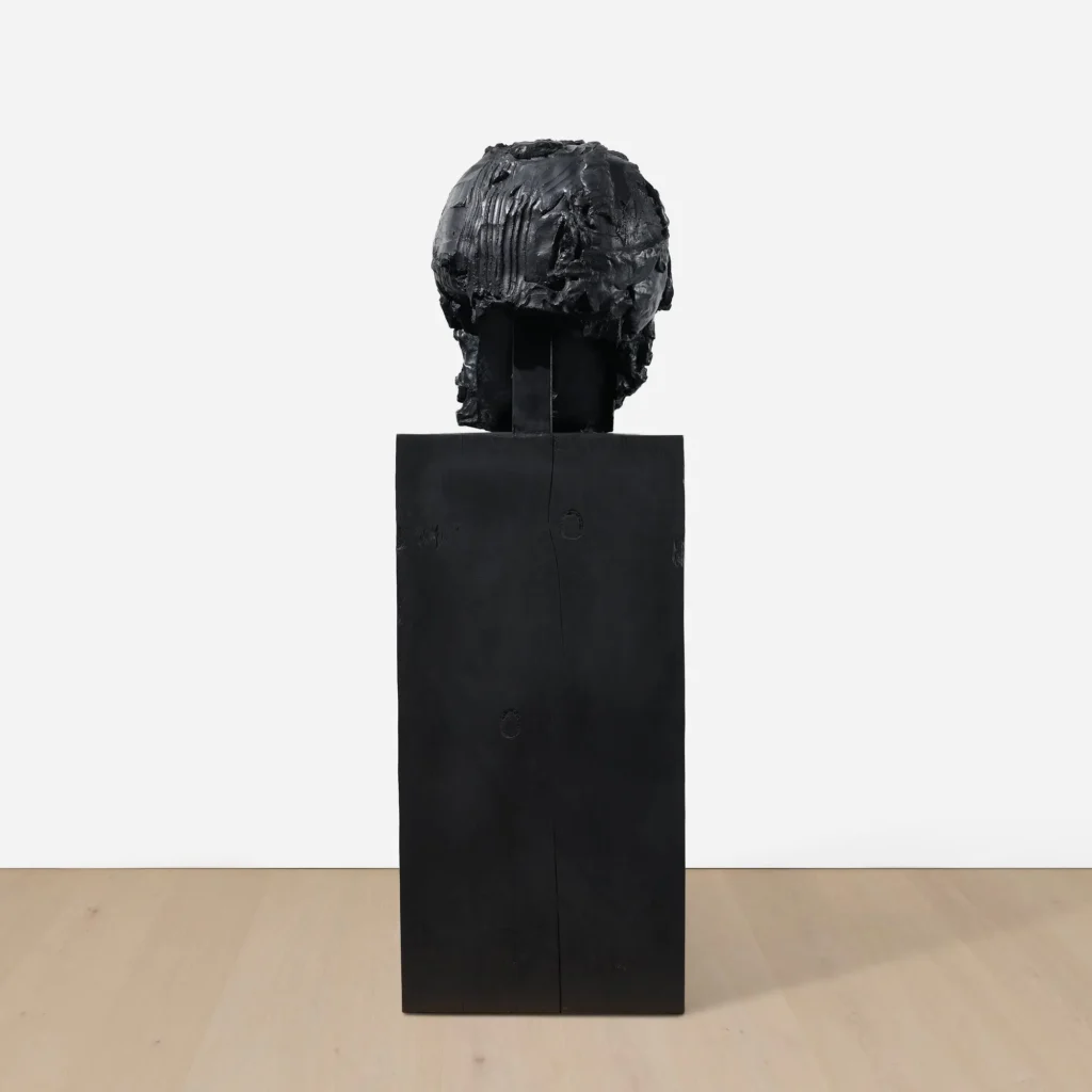 Rear view of a sculptural black skull with deeply grooved textures and an open back structure, displayed on a solid black pedestal against a white gallery wall