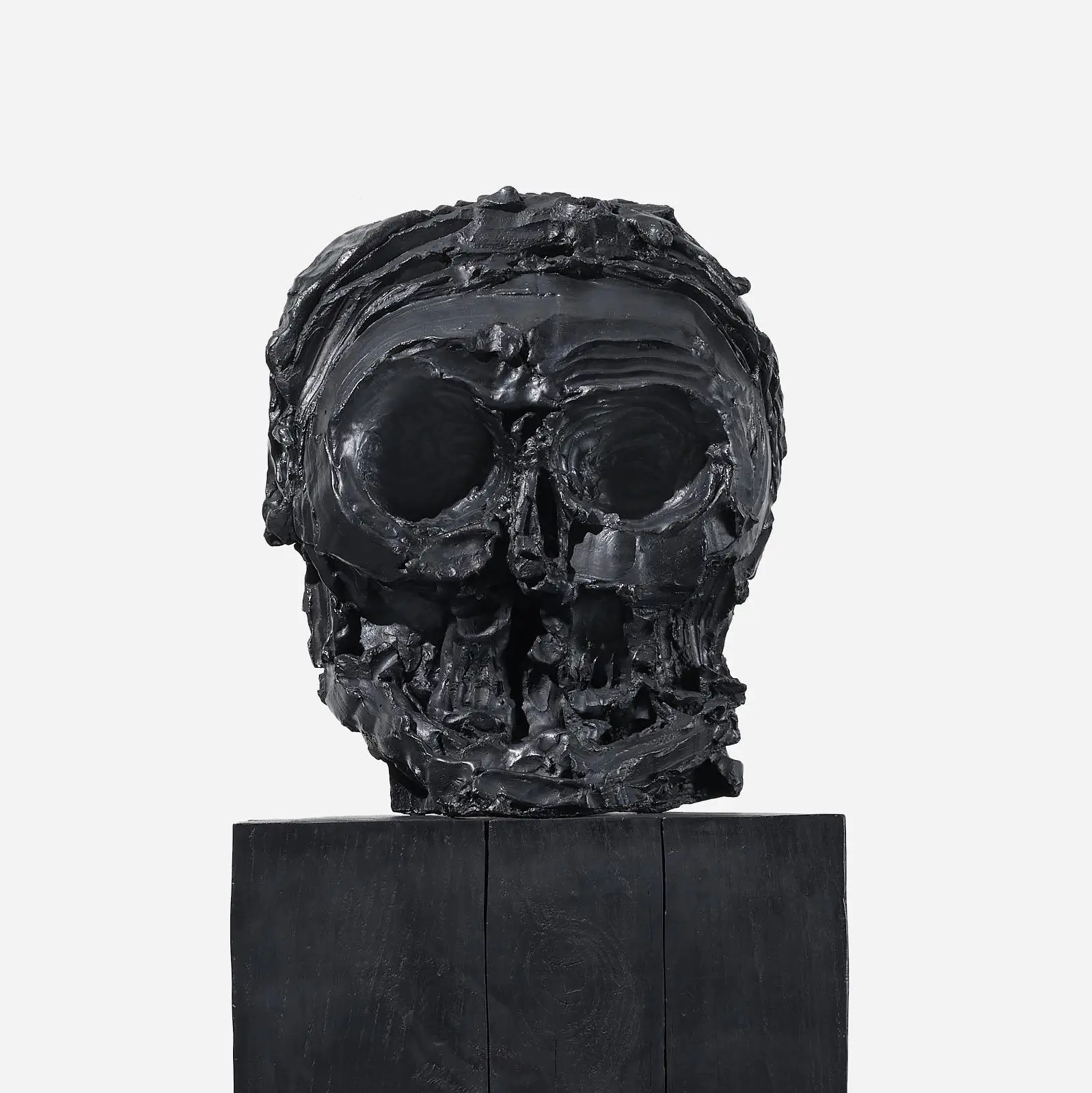 Close-up frontal view of Thomas Houseago's monumental black skull sculpture, revealing heavily layered surfaces, hollow eye sockets, fractured teeth, and expressive hand-worked textures atop a matte-black pedestal.