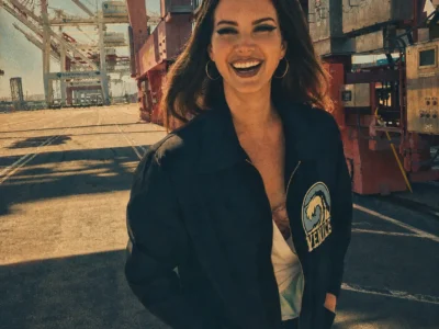 Vintage-inspired portrait of a smiling Lana Del Rey at an industrial shipping port, wearing a black jacket with a Venice wave patch. The image features warm faded tones, heavy 1970s-style film grain, and cargo cranes stretching into the sunlit background