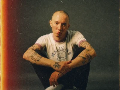 Portrait of Jack Kays seated on the floor against a dark studio backdrop, wearing a white ringer T-shirt, black pants, white socks, and black Converse sneakers. His shaved head, tattoo-covered arms, and direct expression are emphasized by a vintage film treatment featuring heavy grain, warm light leaks, faded colors, dust specks, and subtle scratches that evoke the look of an aged analog photograph
