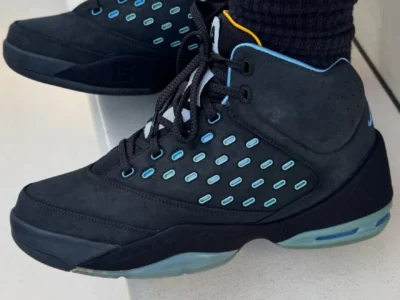 Side view of black Jordan Melo basketball shoe showcasing a nubuck upper with rows of light blue oval eyelet accents across the side panels. The shoes feature black laces, a sculpted black midsole, translucent blue-green Air cushioning, and light blue collar trim with a small yellow detail near the ankle