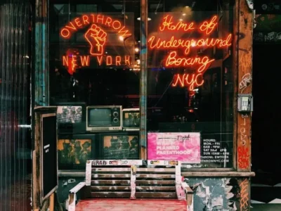 Overthrow Boxing Club storefront in New York featuring neon signs, vintage televisions, graffiti-covered walls, and a weathered red bench outside the entrance