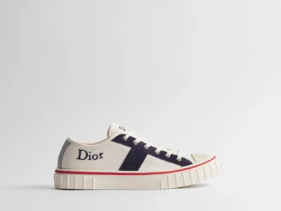 Cream canvas low-top Dior shoe shown in side profile, featuring navy suede side stripes, embroidered Dior logo at the heel, white laces, ribbed rubber toe cap, textured cream rubber sole, and a slim red stripe running around the foxing against a clean white background