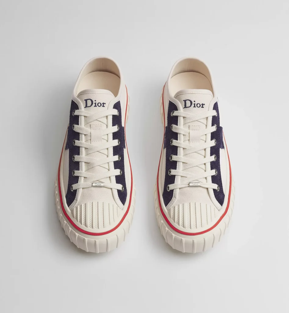 Top-down view of a pair of cream Dior canvas sneakers with navy suede eyelet panels, embroidered Dior logos on the tongues, white flat laces with silver Dior lace tags, ribbed rubber toe caps, and red foxing stripes surrounding the cream platform sole