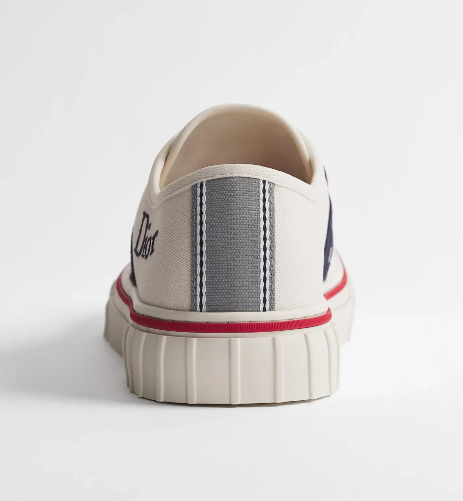 Rear view of the Dior low-top sneaker highlighting the gray woven heel pull tab with white stitched stripes, cream canvas upper, red foxing stripe, and chunky ribbed cream rubber heel against a minimalist white backdrop