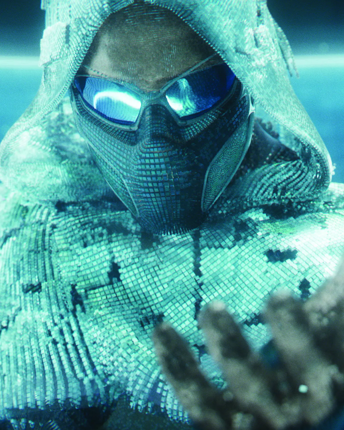 Close-up portrait of a model wearing futuristic Oakley performance sunglasses with mirrored blue lenses, a textured face covering, and a shimmering hooded garment under cool teal lighting.