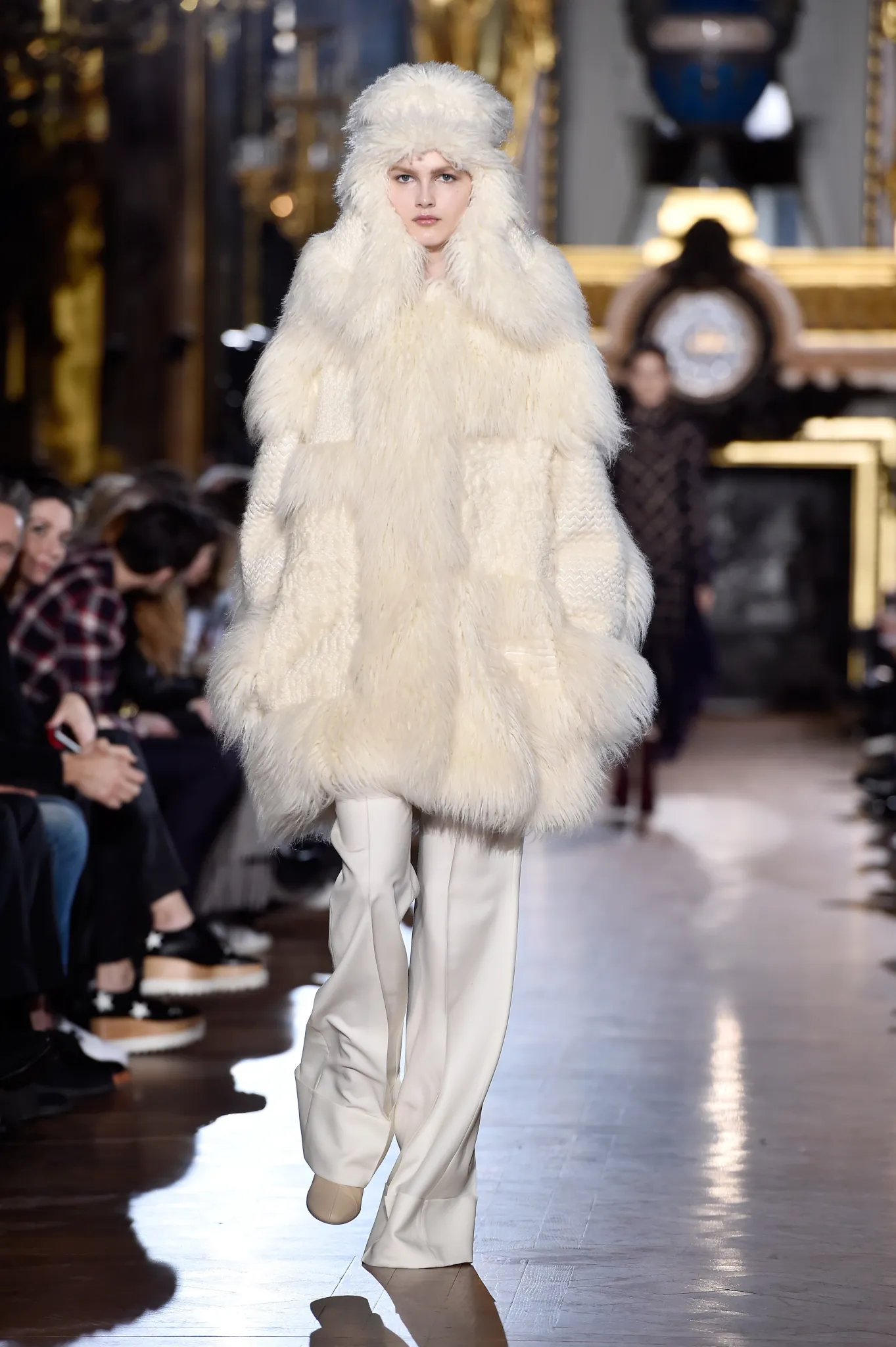 Runway model wearing an oversized ivory faux-fur coat with a matching fur hat, cream wide-leg trousers, and tonal footwear during a luxury fashion show.