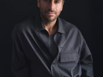 Klitos Teklos wearing a charcoal gray utility-style overshirt with a large flap chest pocket, layered over a matching knit top and coordinated belted trousers. He has short light brown hair, light stubble, and a calm expression while facing the camera against a dark black background with soft, directional lighting that emphasizes the minimalist tailoring and clean sil