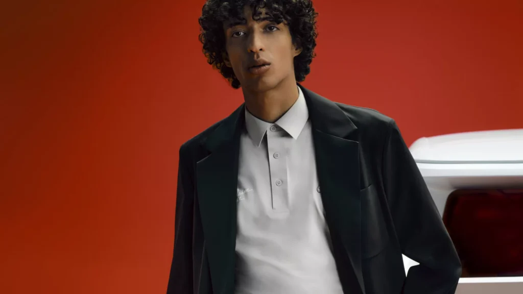 Model wearing a black tailored jacket over a white Alpine x Lacoste polo shirt, posed beside the rear of the Alpine A290 Rallye with a bold red background