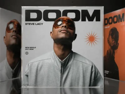 Editorial-style promotional artwork for Steve Lacy's "DOOM" single, featuring a central portrait of the artist in oversized amber-tinted sunglasses and a light gray perforated zip-up jacket, surrounded by multiple stylized album covers in a Cover Flow-inspired display with bold black typography and orange graphic accent