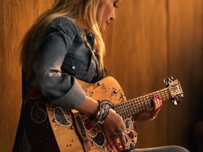 Dolly Parton in a denim shirt and jeans sits on a wooden cabinet while playing an embellished acoustic guitar decorated with rhinestones and paisley-inspired patterns. Warm golden lighting highlights her profile, layered bracelets, rings, and red nail polish against a softly blurred wood-paneled background, creating an intimate, high-resolution portrait