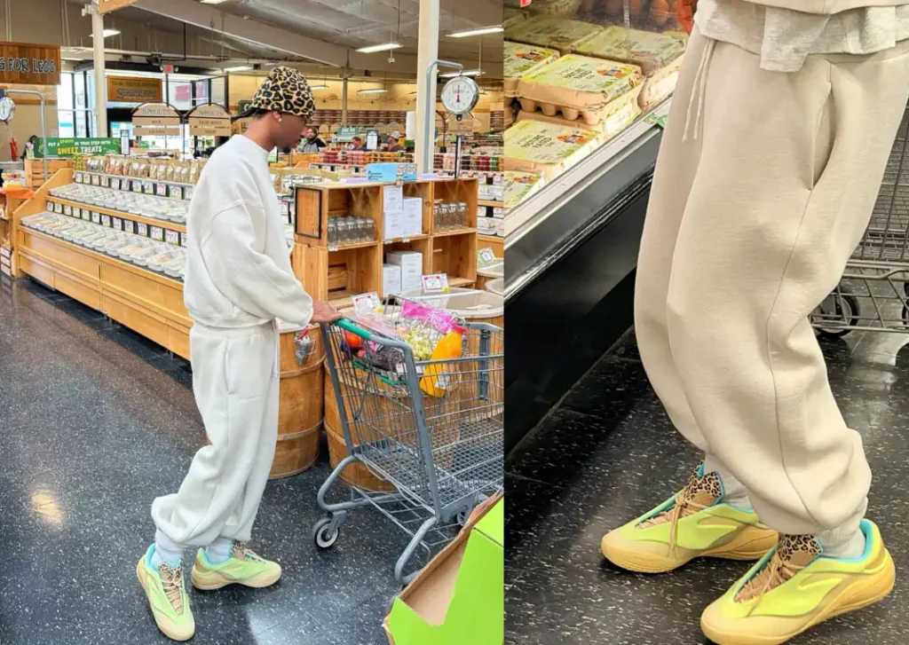 A person in a grocery store wears a leopard print hat, a loose white sweatshirt, matching sweatpants, and yellow sneakers with blue and leopard print accents. They push a shopping cart near shelves of produce and eggs.