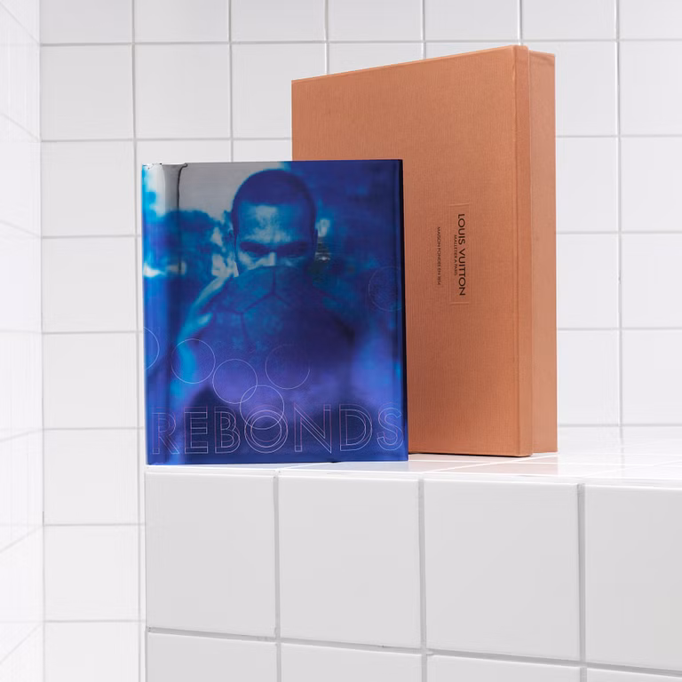 The Louis Vuitton Rebonds collector's book displayed alongside its signature orange presentation box on a white tiled surface. The book features a blue-toned cover with a portrait partially obscured by a soccer ball, reflecting the publication's intersection of football, fashion, and contemporary culture.
