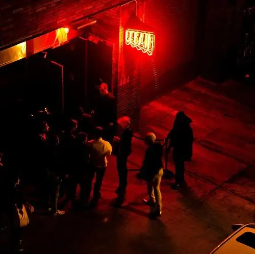 Nighttime street scene showing a small crowd gathered outside a venue beneath a glowing red neon sign, viewed from an elevated angle with dramatic lighting.