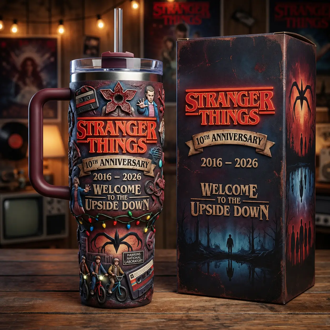 A large insulated Stranger Things 10th Anniversary travel tumbler with a matching handle and straw stands beside its collector's gift box. The cup is decorated with raised artwork featuring Eleven, the Demogorgon, cassette tapes, bicycles, Christmas lights, and the Welcome to the Upside Down theme, set against a retro entertainment room backdrop.