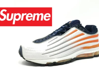 White and navy Supreme x Nike running shoe with bold orange side stripes, sculpted ribbed overlays, black outsole, and a sleek aerodynamic silhouette against a clean white background, with a partial Supreme box logo view on the left