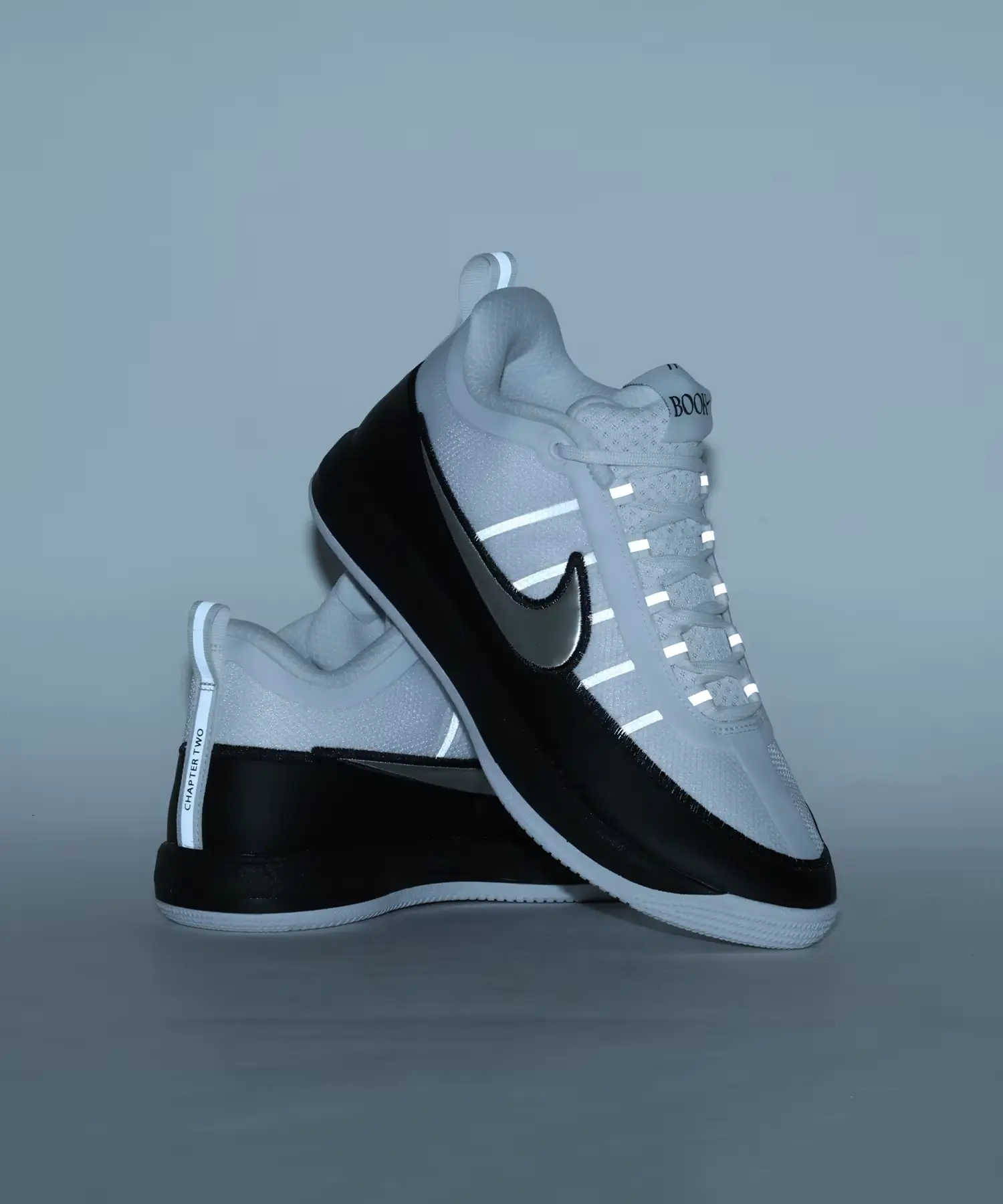 Pair of Nike BOOK Chapter Two sneakers stacked in a studio with reflective lace loops and heel pull tabs illuminated, highlighting the white mesh upper, metallic silver Swoosh, and black mudguard.