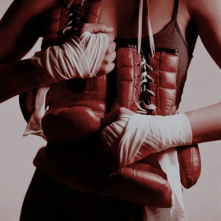 Person with white hand wraps holding a pair of worn brown leather boxing boots against a dark athletic tank top, creating a dramatic portrait centered on boxing heritage and training.
