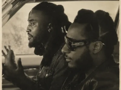 Vintage-inspired 1970s monochrome portrait of Shaboozey and Leon Bridges seated inside a classic car, viewed through the open window. The driver gestures while speaking, and the passenger wears oversized sunglasses and a leather jacket. The image features a soft off-white sepia tone, heavy film grain, faded contrast, and worn photo border