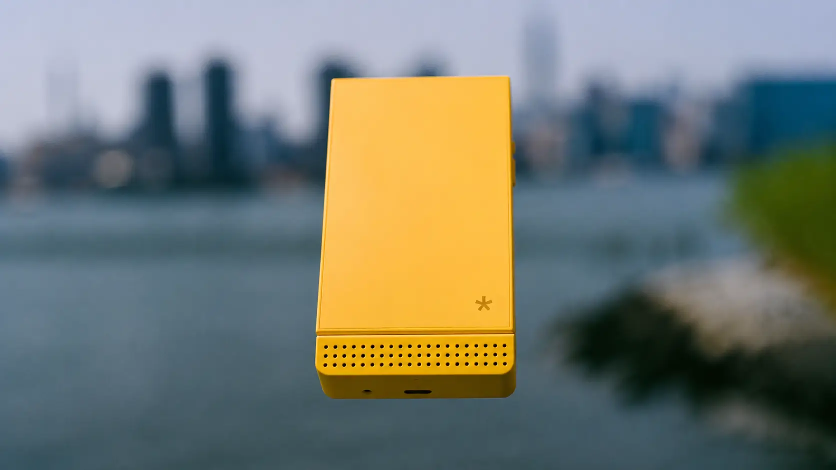 Bright yellow Light Phone shown upright against a softly blurred waterfront skyline, highlighting its minimalist front design with a perforated speaker grille and clean industrial aesthetic.