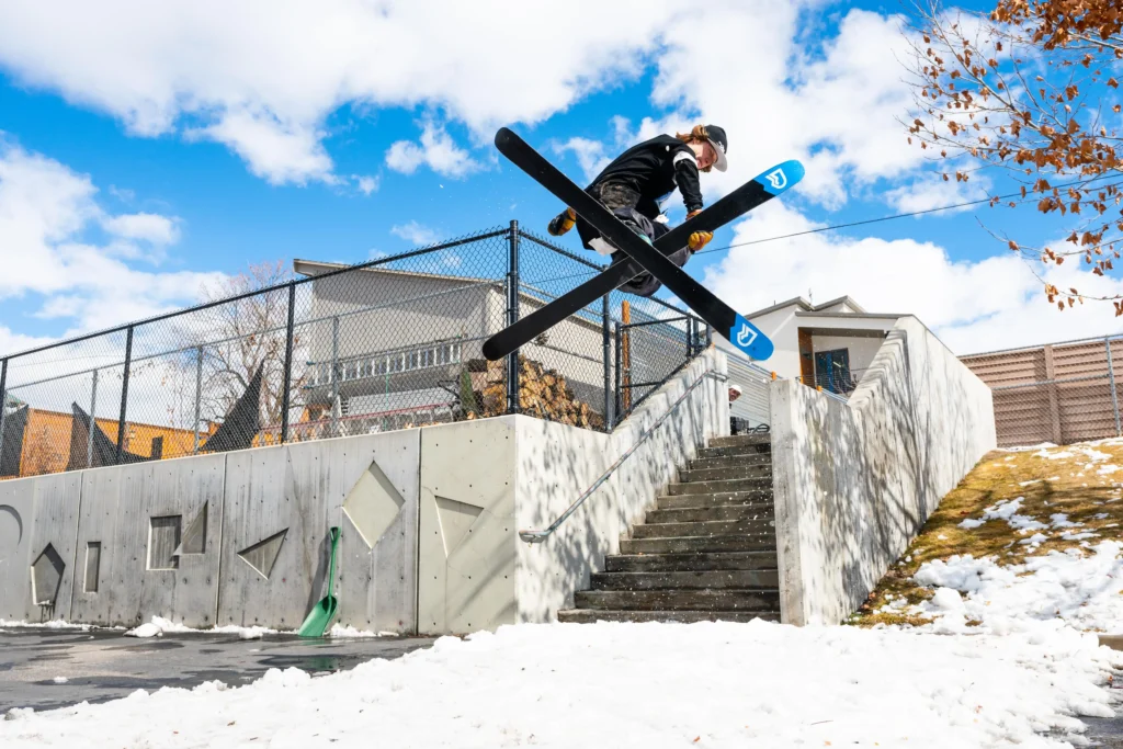 Freestyle skier launches high above a concrete stair set in an urban neighborhood, crossing skis mid-air over a handrail with blue skies, patchy snow, and modern homes in the background