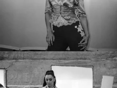 Black-and-white behind-the-scenes photo of Charli xcx seated on film equipment at an outdoor production set, surrounded by crew members, lighting rigs, and camera gear